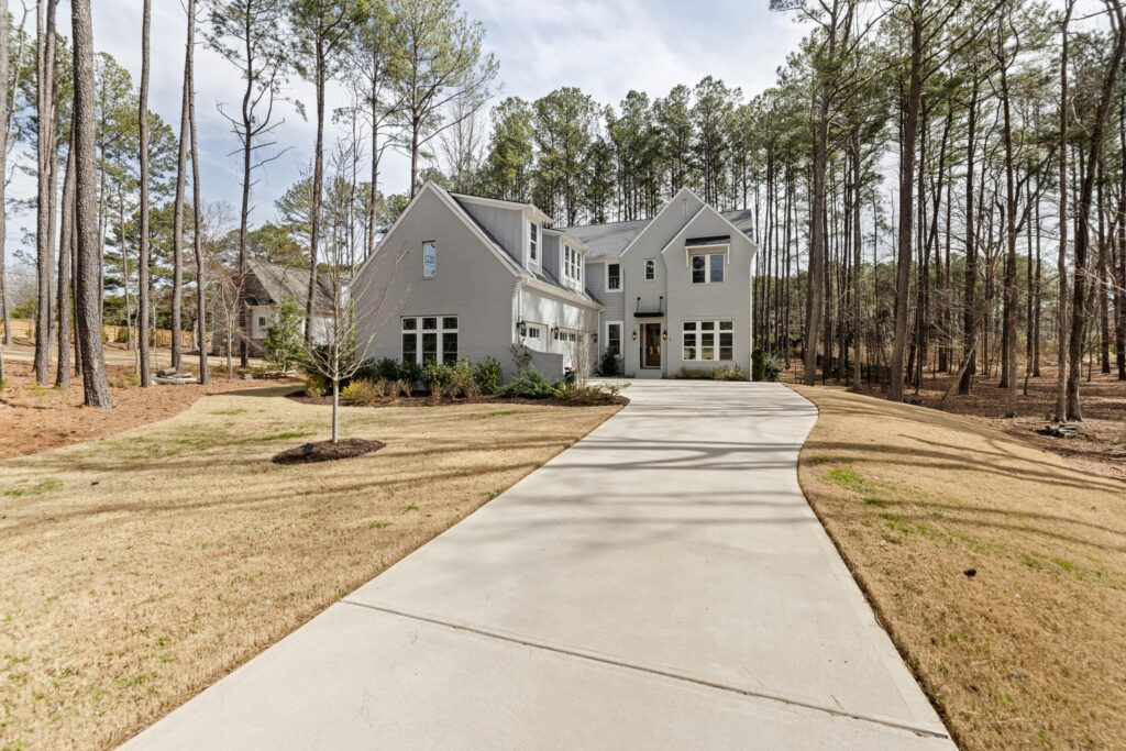 Gray two-story house with multiple gabled roofs at the end of a curved concrete driveway, surrounded by tall pine trees amid a sparse lawn.